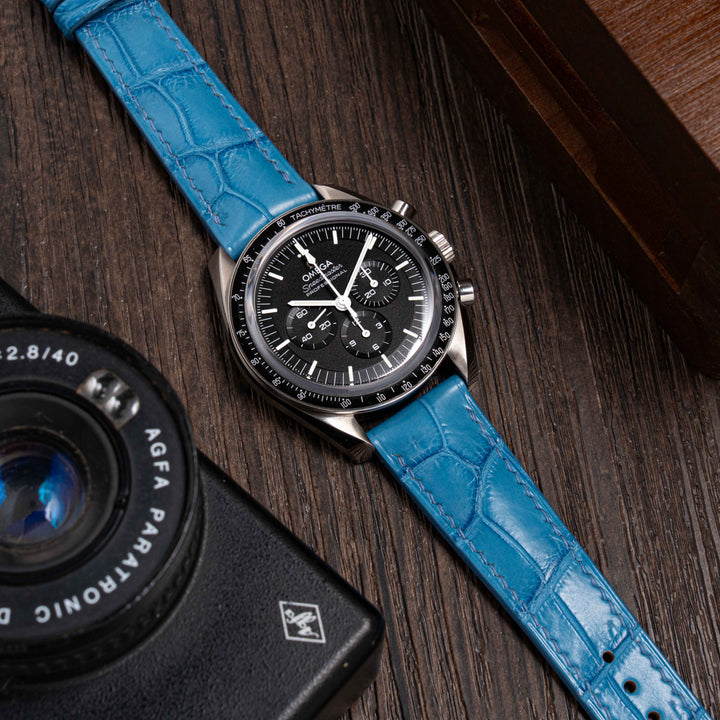 Watch with blue strap and black dial Omega speedmaster 
on a wooden surface next to an Agfa Parrotonic camera.