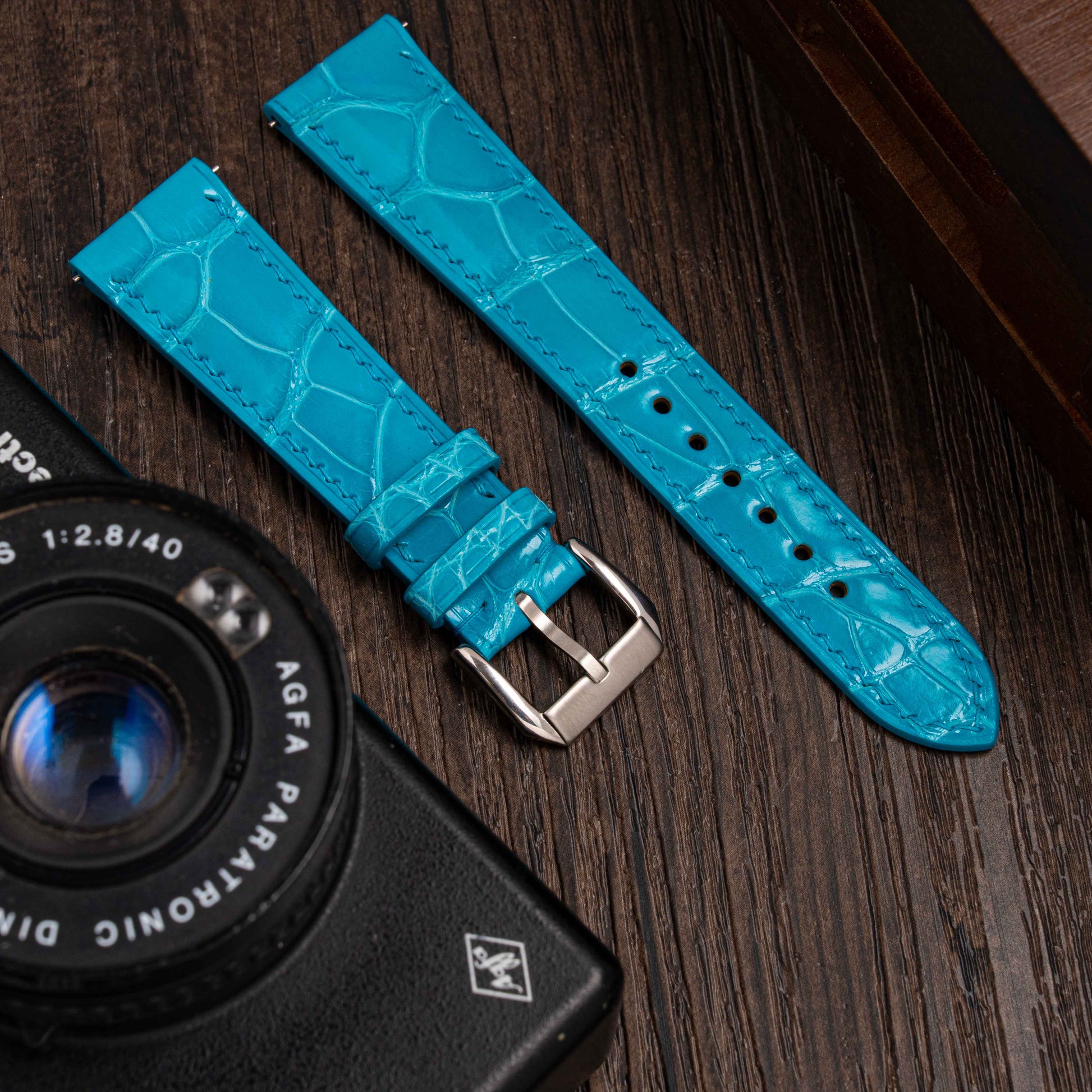 Two blue watch straps on a wooden surface with an Agfa camera lens.