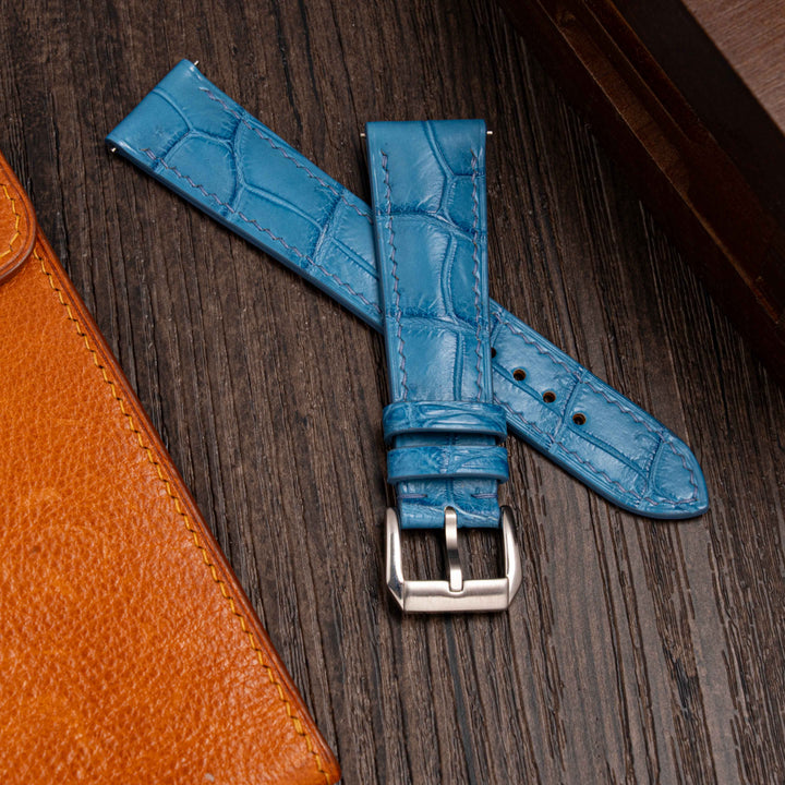 Blue crocodile-patterned watch strap with silver buckle on a wooden surface